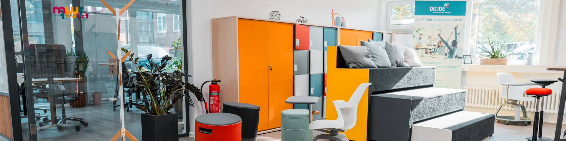KOMMA Header MWF Ein moderner Büroraum mit farbenfrohen Schränken, einem abgestuften Sitzbereich, verschiedenen ergonomischen Stühlen, Pflanzen, einem Feuerlöscher und großen Fenstern, die natürliches Licht hereinlassen.