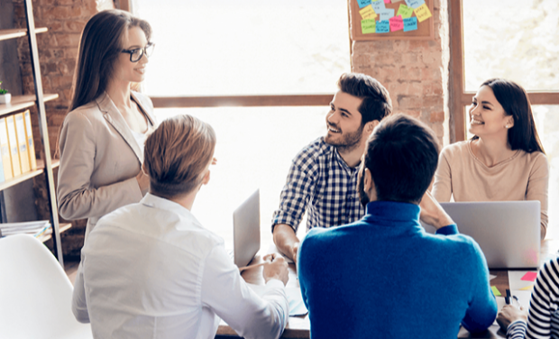 Fünf Personen sitzen in einem modernen Büro um einen Tisch herum. Eine Frau steht und spricht, während die anderen, die mit Laptops und Notebooks sitzen, zuhören und lächeln. An einem Fenster hinter ihnen sind Klebezettel zu sehen.