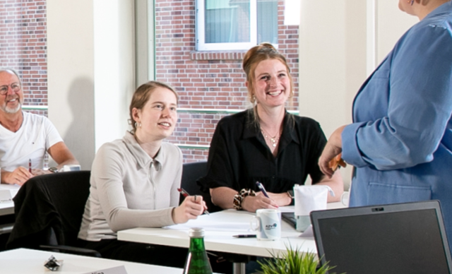 Zwei Frauen sitzen an einem Tisch, lächeln und machen sich Notizen, während sie einer stehenden Person in einer hellblauen Jacke zuhören. Auf dem Tisch stehen Büromaterial und eine Pflanze. Im Hintergrund sind ein Fenster und eine Backsteinmauer zu sehen.