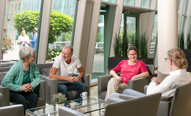 Vier ältere Erwachsene sitzen und unterhalten sich um einen gläsernen Couchtisch in einem hellen, modernen Aufenthaltsraum von Fortbildung KOMMA Bordesholm. Durch die großen Fenster ist Grün zu sehen, im Hintergrund stehen zwei Personen in der Nähe des Eingangs.