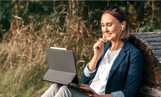 Eine Frau sitzt im Freien an einem Baumstamm und lächelt, während sie ein Tablet benutzt und Kopfhörer trägt, was darauf hindeutet, dass sie ein Videogespräch führt oder etwas hört. Im Hintergrund sind hohes Gras und Bäume zu sehen.