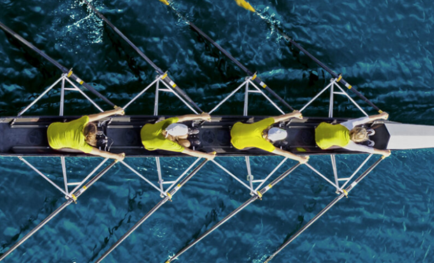 Luftaufnahme von vier Ruderern in gelben Hemden und weißen Mützen, die gemeinsam in einem Komma-Rennboot auf dem klaren blauen Wasser rudern.