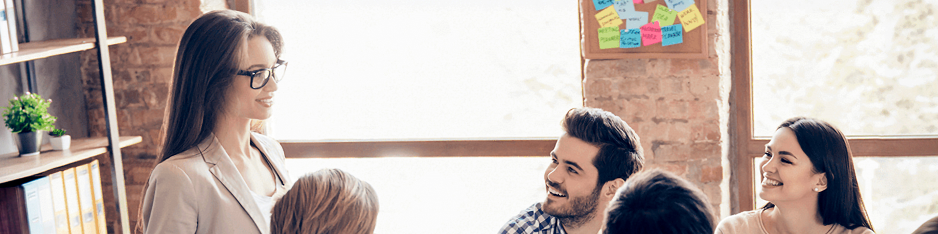 Eine Gruppe von Menschen sitzt um einen Tisch in einem hellen Büro, lächelt und unterhält sich während einer Fortbildungssitzung. Eine Frau steht auf und spricht zu der Gruppe, während im Hintergrund bunte Klebezettel auf einer Tafel zu sehen sind.