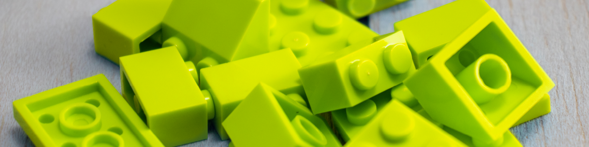 KOMMA Header Modul Eine Nahaufnahme von mehreren verstreuten gelb-grünen Plastikbausteinen auf einer hellen Holzoberfläche.