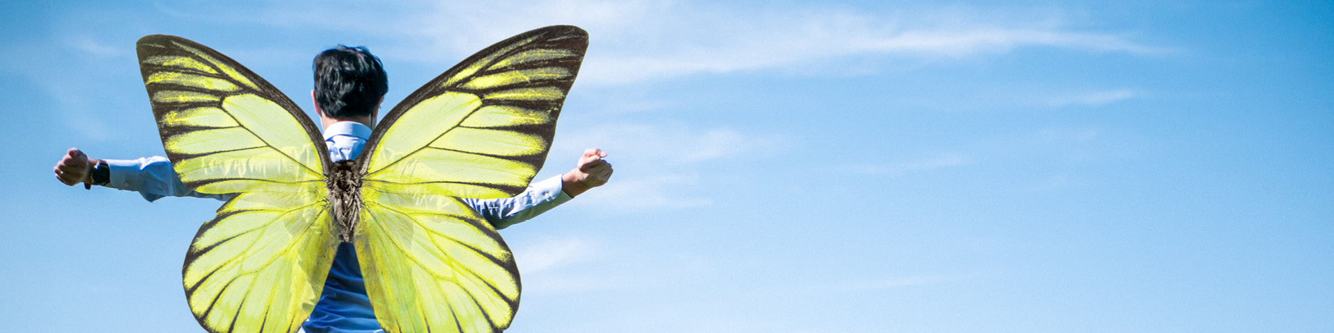 Eine Person steht mit ausgestreckten Armen vor einem blauen Himmel und hat große, grüne Schmetterlingsflügel auf dem Rücken.